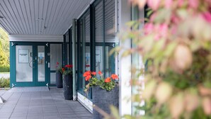 Une entrée d’immeuble avec un porche abrité, une porte avec une enseigne et des plantes en pot ornées de fleurs rouges.