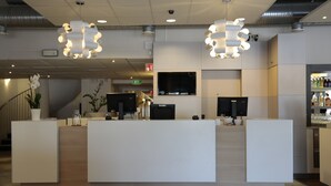 Un espace d’accueil au design moderne, comprenant un comptoir de réception, deux grands luminaires suspendus, un téléviseur à écran plat et un bar avec des bouteilles exposées.