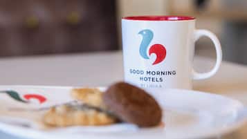 A white mug with a logo and text on a table with a plate of food and a brown chair in the background.