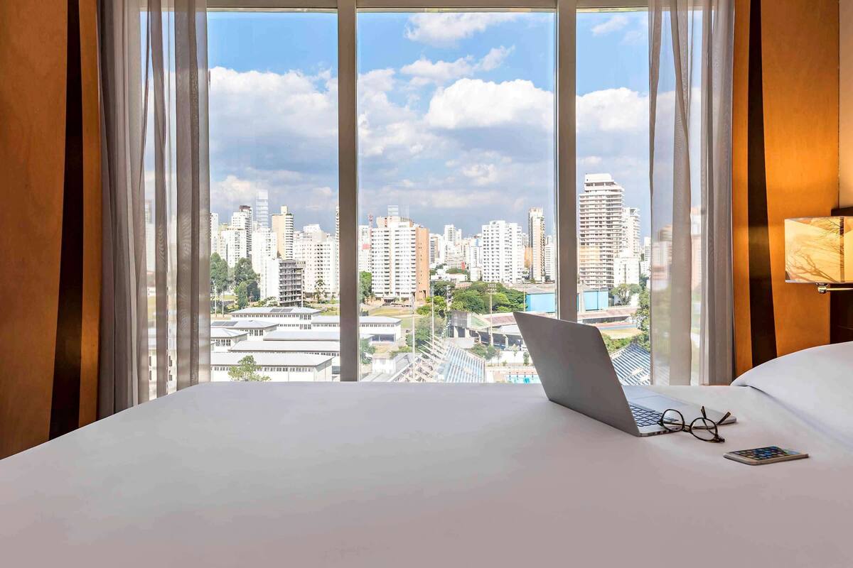 Habitación de hotel con un ventanal grande que ofrece vista a la ciudad, una cama, una laptop y un par de gafas sobre la cama.
