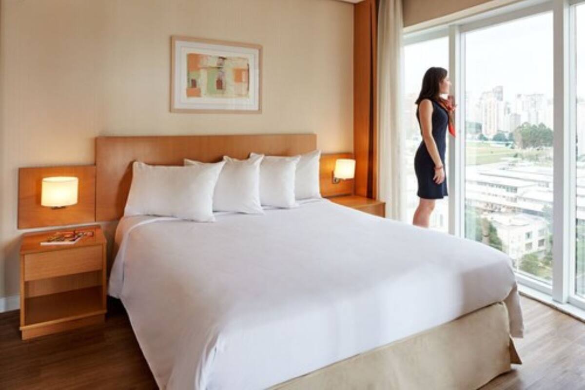 Habitación de hotel con una cama grande, dos mesitas de noche con lámparas, un cuadro en la pared y vistas de edificios a través de la ventana.