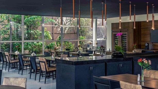 A modern restaurant with a central bar area, large windows offering views of greenery, and contemporary lighting fixtures. - Lummina Ibirapuera São Paulo (Sao Paulo)