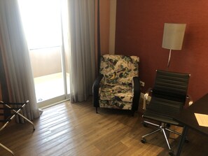 Een kamer met een houten vloer, een fauteuil met bloemenprint, een zwarte bureaustoel, een bureau en een lamp.