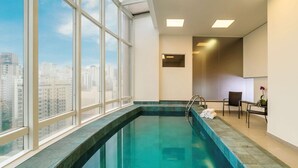 A modern indoor pool with clear glass walls, a view of the cityscape, and a seating area with chairs and a small table. - Lummina Ibirapuera São Paulo (Sao Paulo)