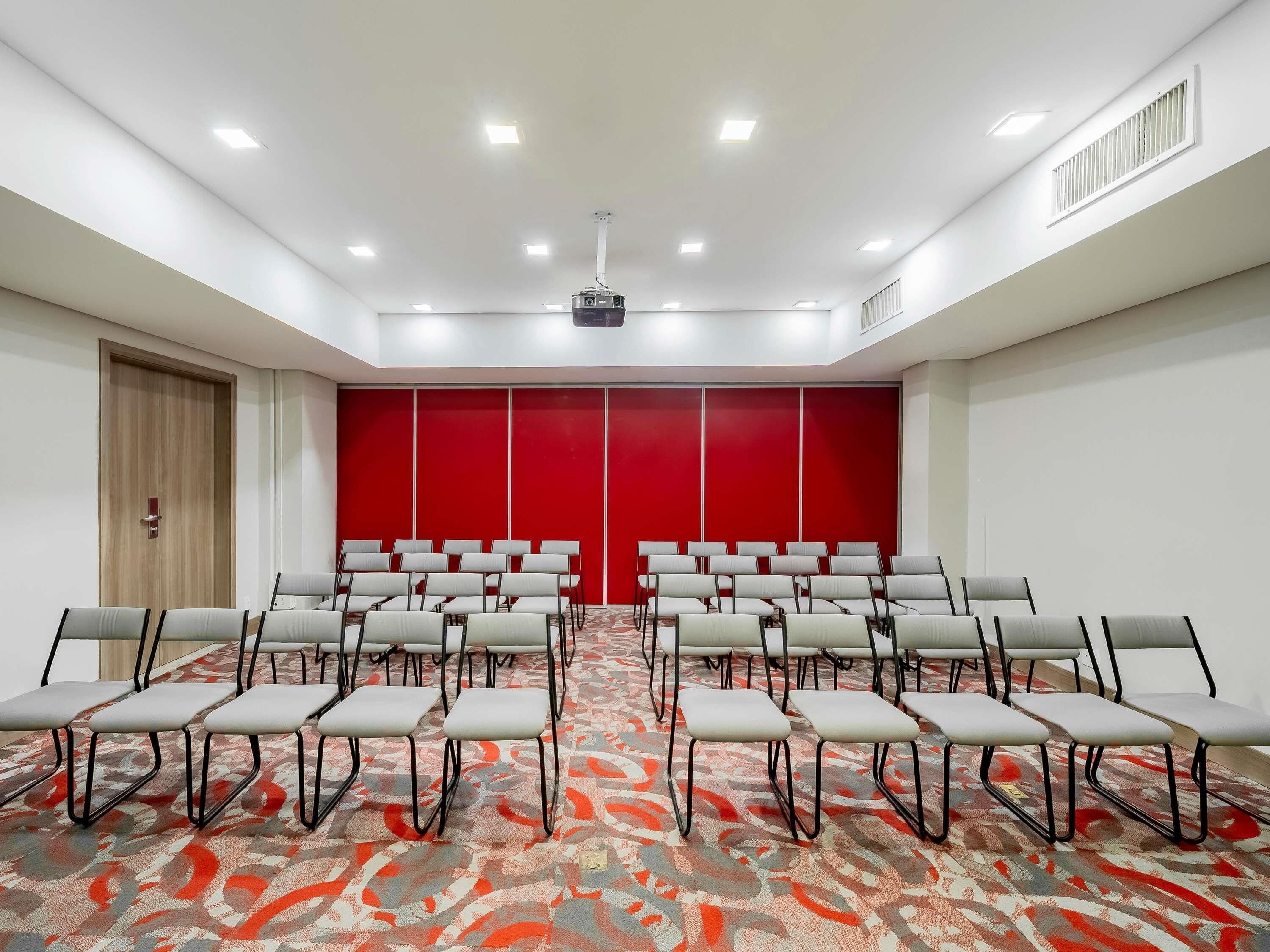 Un salón de conferencias con filas de sillas, un proyector y una pared de acento rojo.