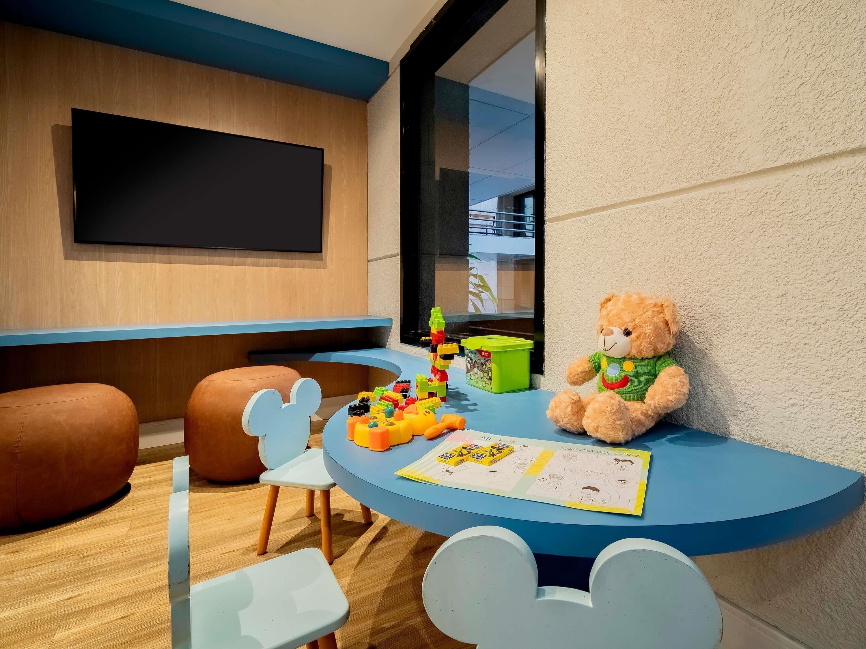 Uma área de recreação infantil com um urso de pelúcia grande, blocos de construção coloridos e uma mesa em formato do Mickey Mouse.