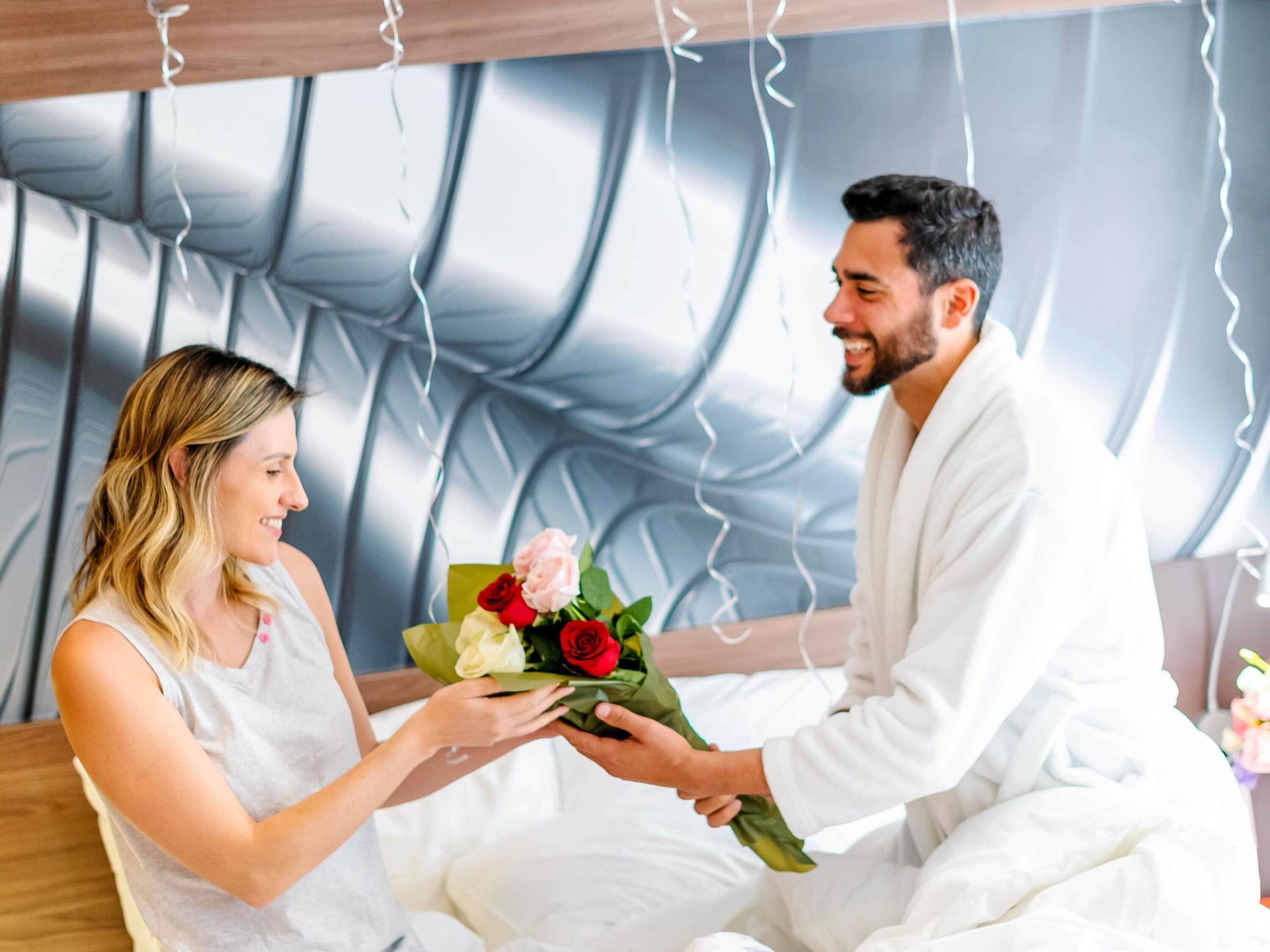 A person in a white bathrobe presenting a bouquet of flowers to another individual in a white sleeveless top.