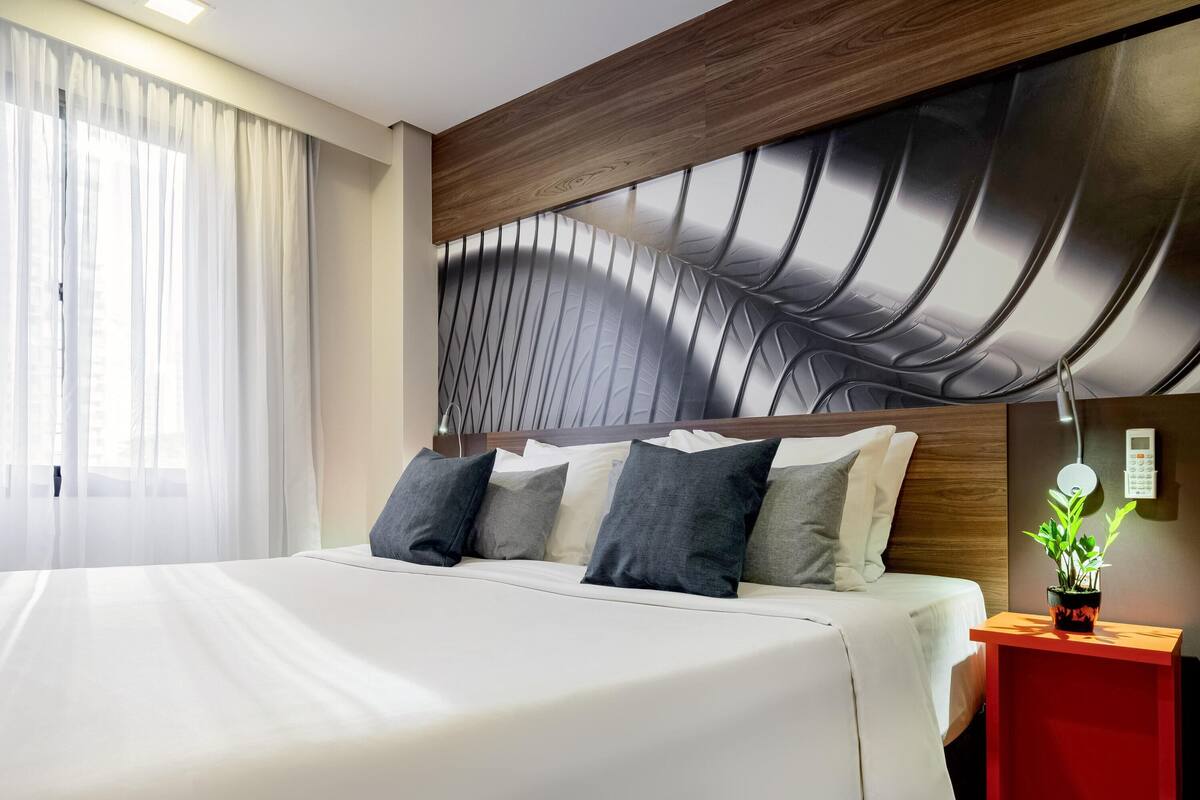 Habitación de hotel con una cama grande, un cabecero moderno, una mesita de noche con una mesita auxiliar roja y una planta en maceta.