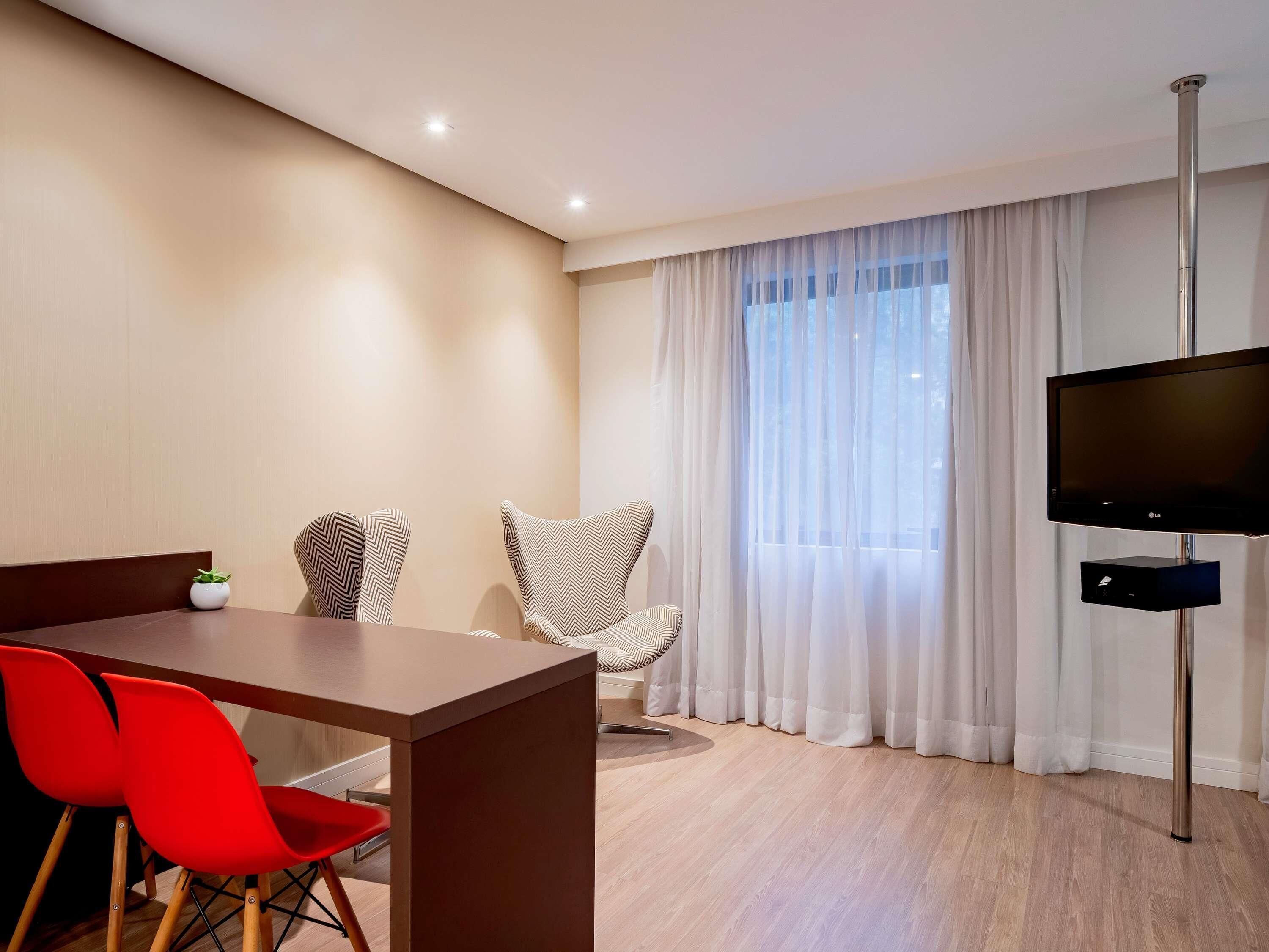 Ein stilvolles Zimmer mit Holztisch, roten Stühlen, einem Fernseher und einem großen Fenster mit leichtem Vorhang.