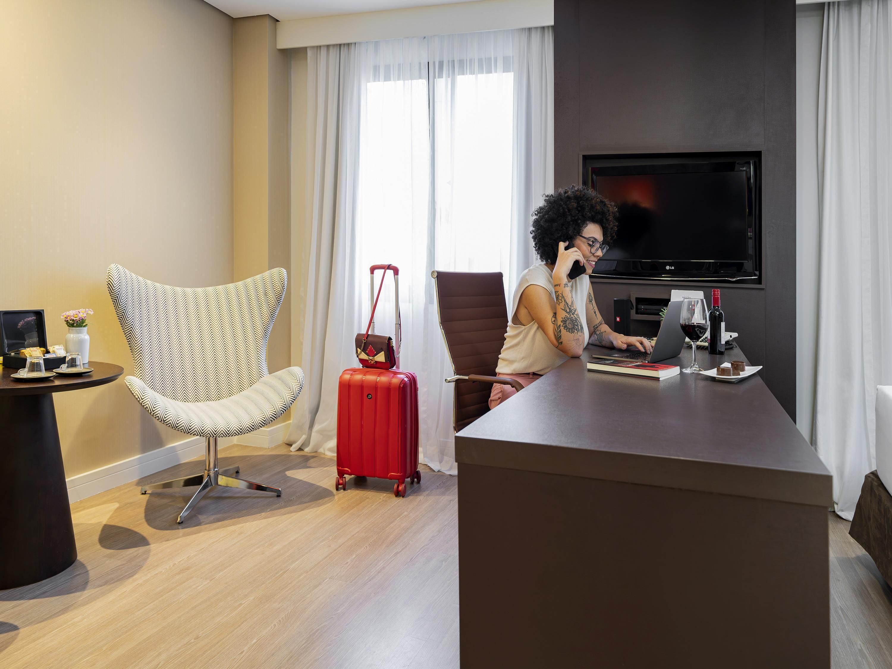A hotel room with a modern chair, a desk with a laptop, a red suitcase, and a TV.