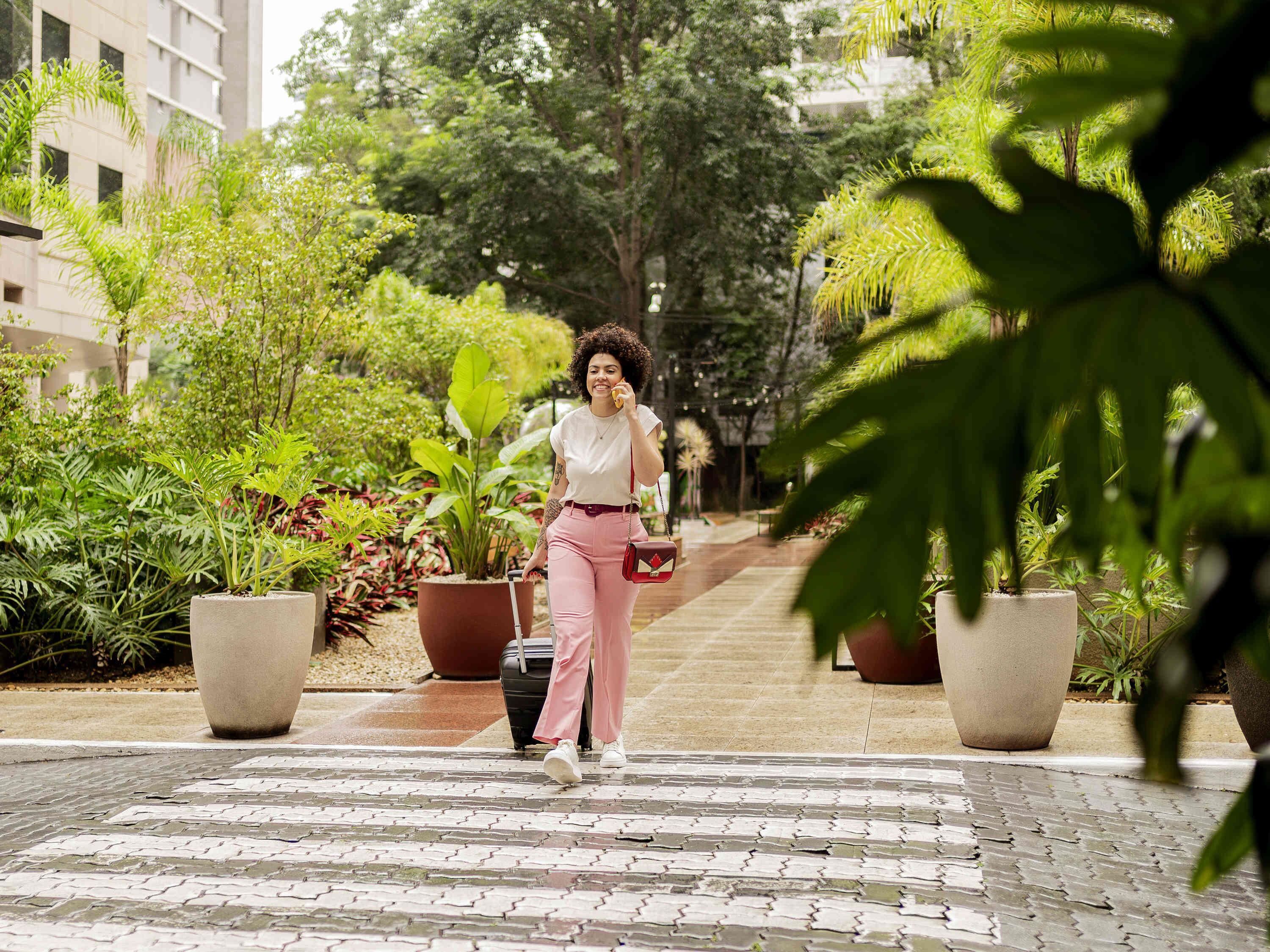 A person walking with a suitcase in a paved outdoor area with potted plants and a building in the background.
