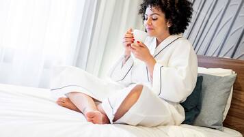 Una persona sentada en una cama con un libro, vestida con batas blancas, en una habitación con cabecera de madera y una ventana con cortinas.