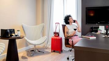 A hotel room with a large flat-screen TV, a swivel chair, a small table with a coffee maker, a red suitcase, and a desk with a laptop and a bottle of wine.
