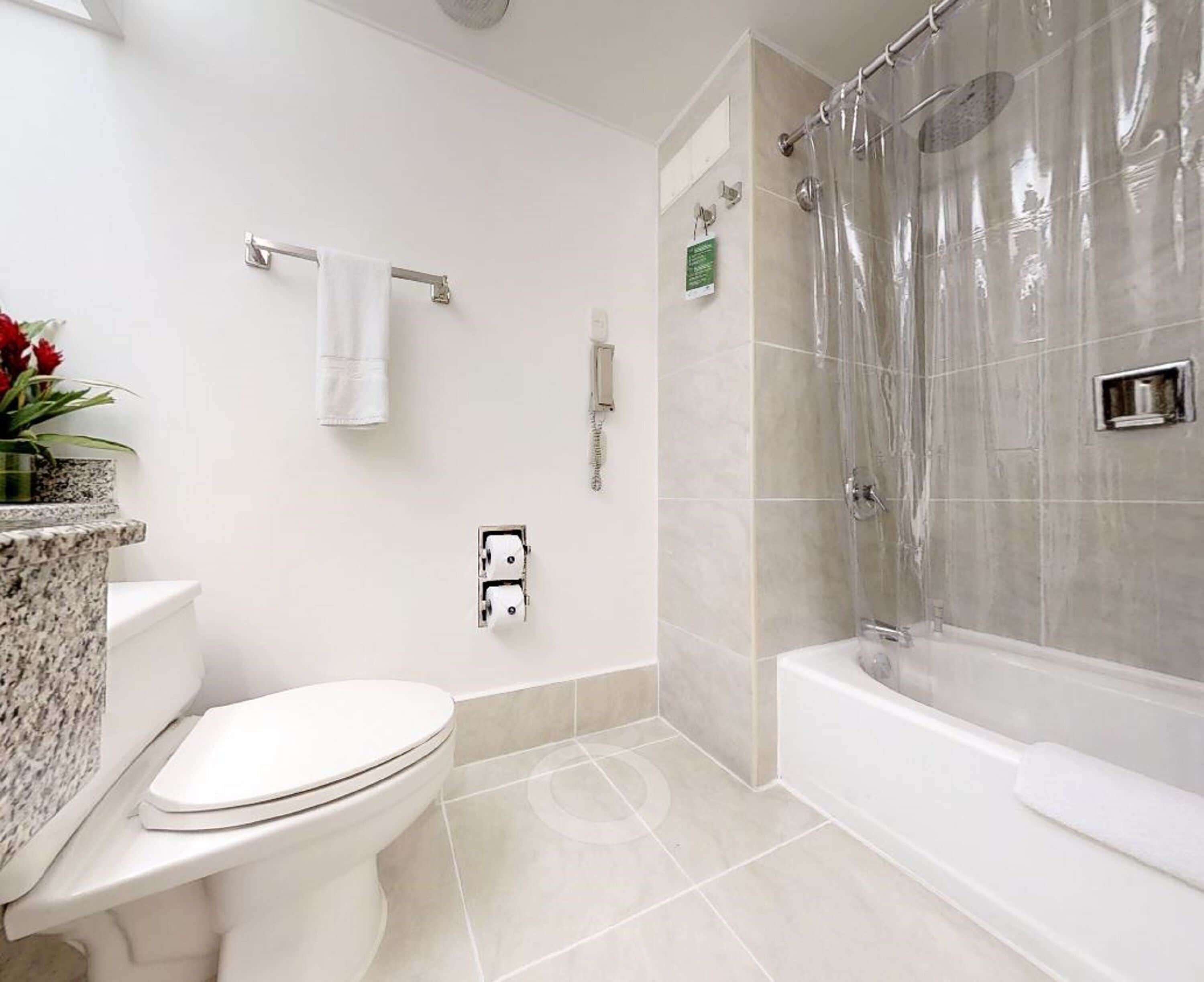 A bathroom with a toilet, a shower with a transparent curtain, and a marble countertop with a plant.