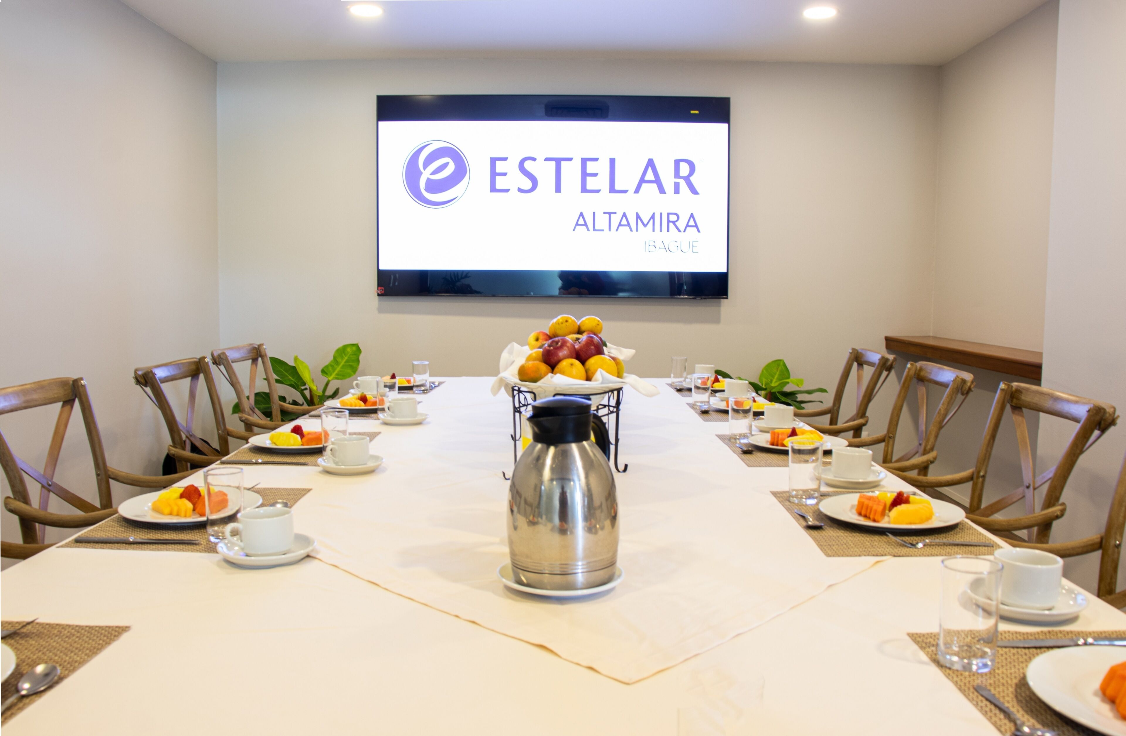 Ein Konferenzraum mit einem langen Tisch, Stühlen, einer Kaffeemaschine und Obst auf Tellern. Ein Fernsehbildschirm zeigt den Namen des Hotels.