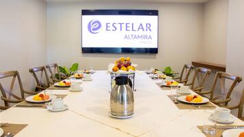 Ein Konferenzraum mit einem langen Tisch, Stühlen, einer Kaffeemaschine und Obst auf Tellern. Ein Fernsehbildschirm zeigt den Namen des Hotels.