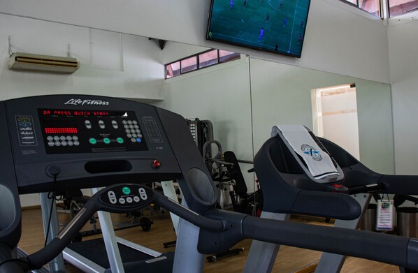A gym with a treadmill and a stationary bike, a control panel, and a TV screen displaying a soccer game. - Hotel Estelar Altamira (Ibagué)