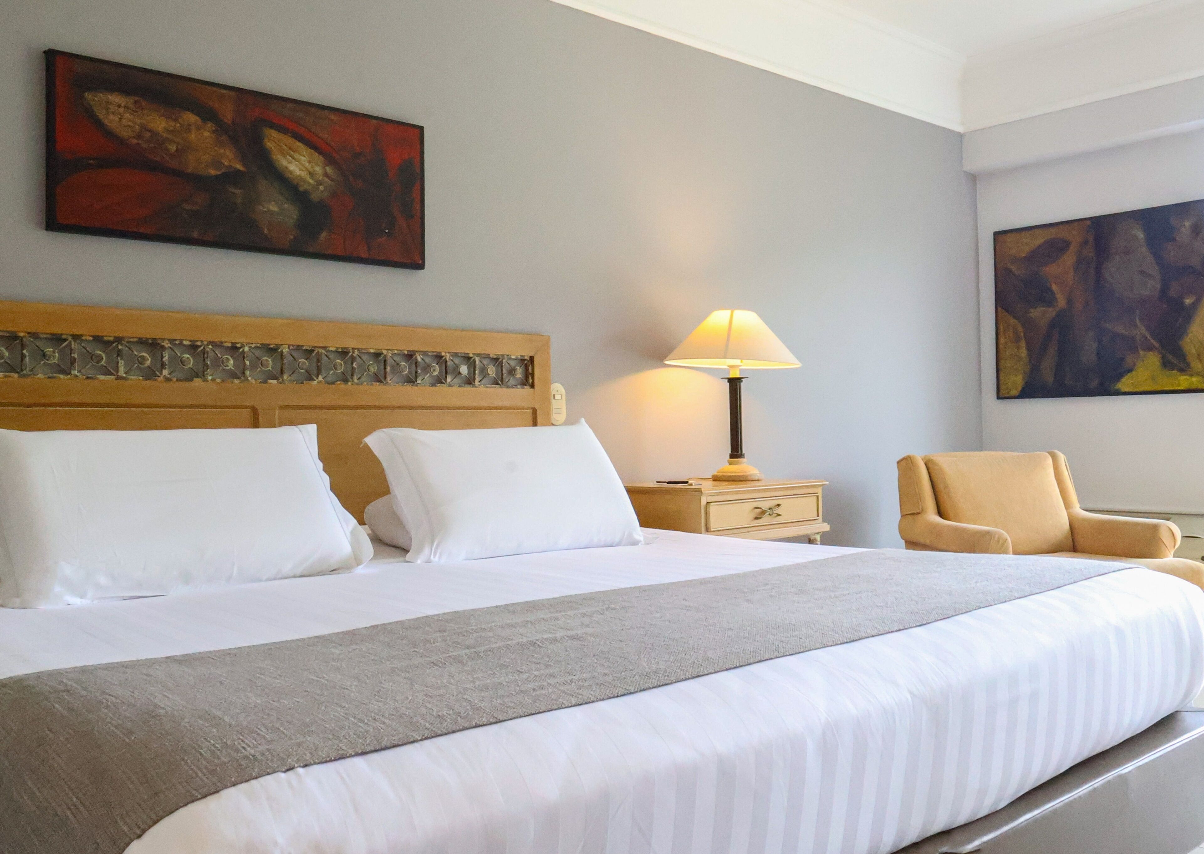 A hotel room with a large bed, a nightstand with a lamp, a chair, and two pieces of wall art.
