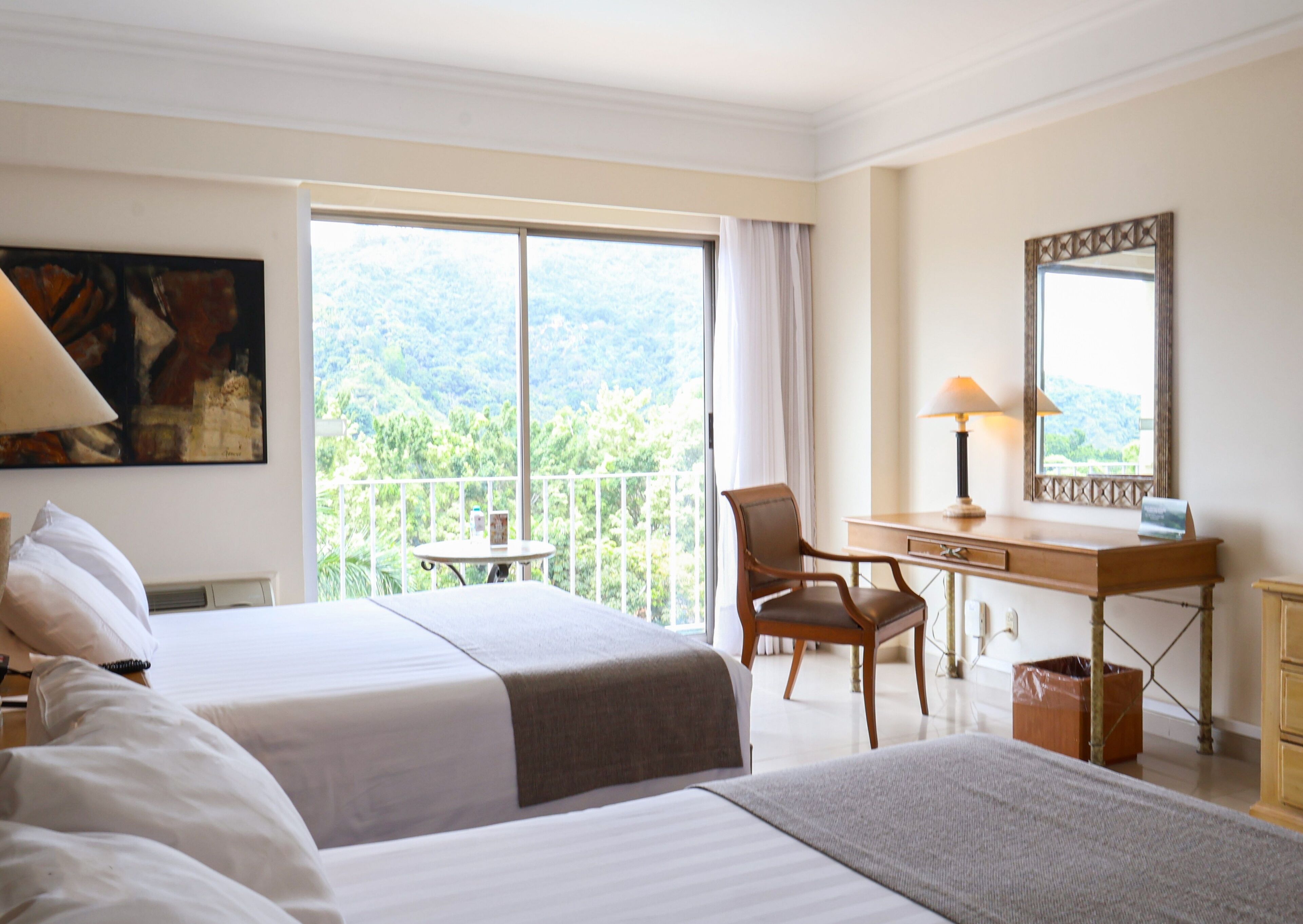 A hotel room with two beds, a desk, a chair, a window with a view of greenery, and a painting on the wall.