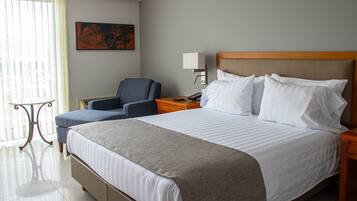 A hotel room with a large bed, a chair, a nightstand, a lamp, and a painting on the wall.