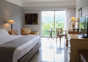 Habitación de hotel con cama, dos sillas, escritorio con televisor y balcón con vistas a la vegetación.
