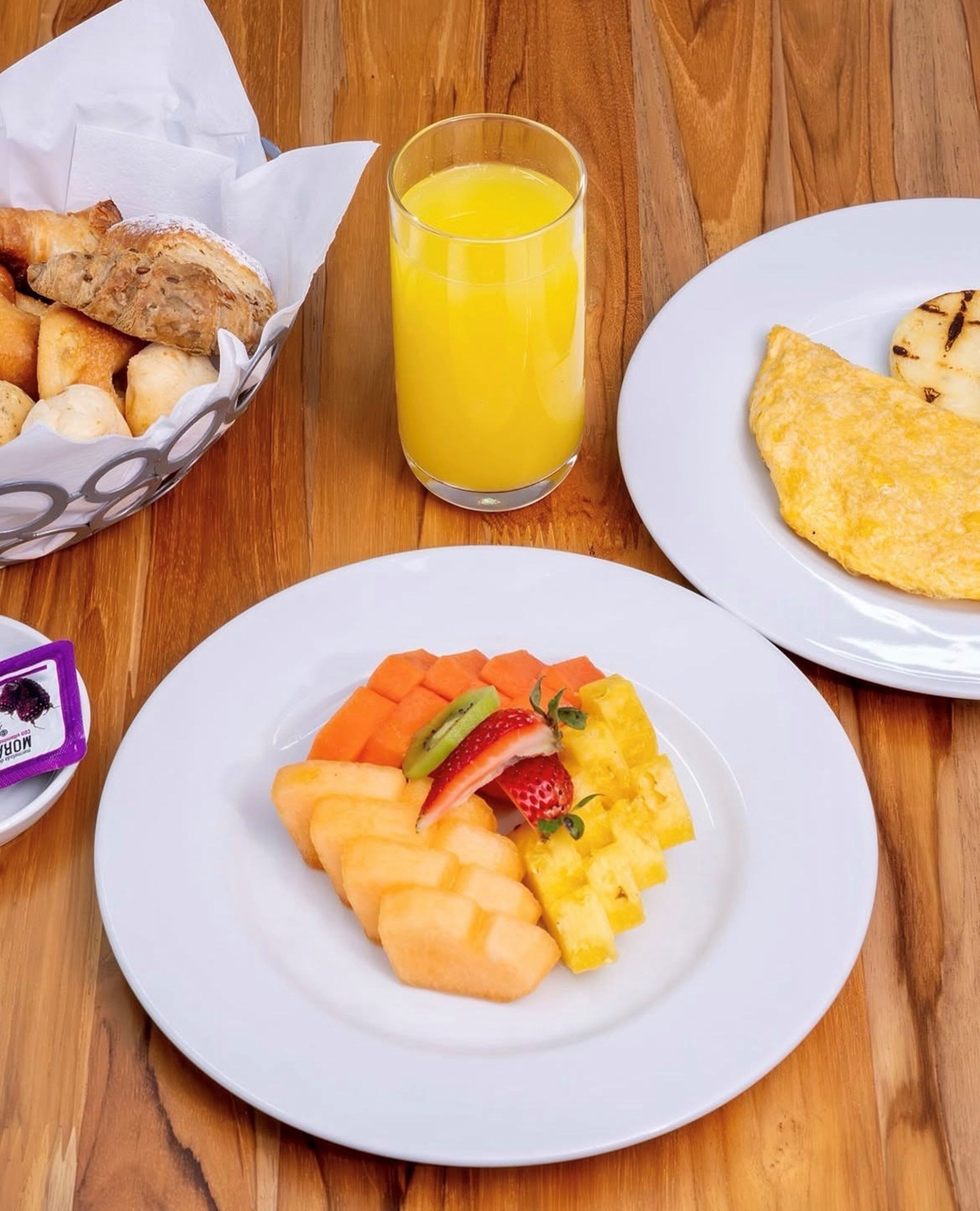 A breakfast setting with a plate of fruit, a glass of orange juice, a basket of bread, and a plate with an omelette.