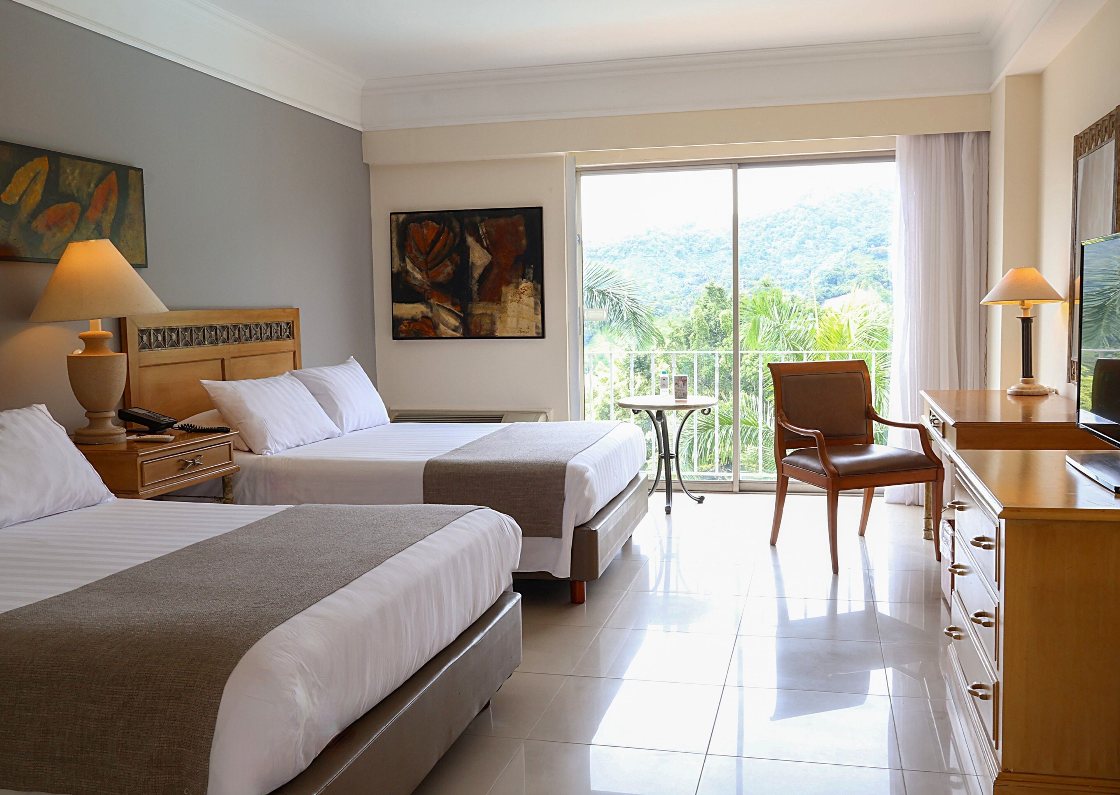 A hotel room with two beds, a desk, a chair, a TV, a balcony with a view of greenery, and artwork on the walls.