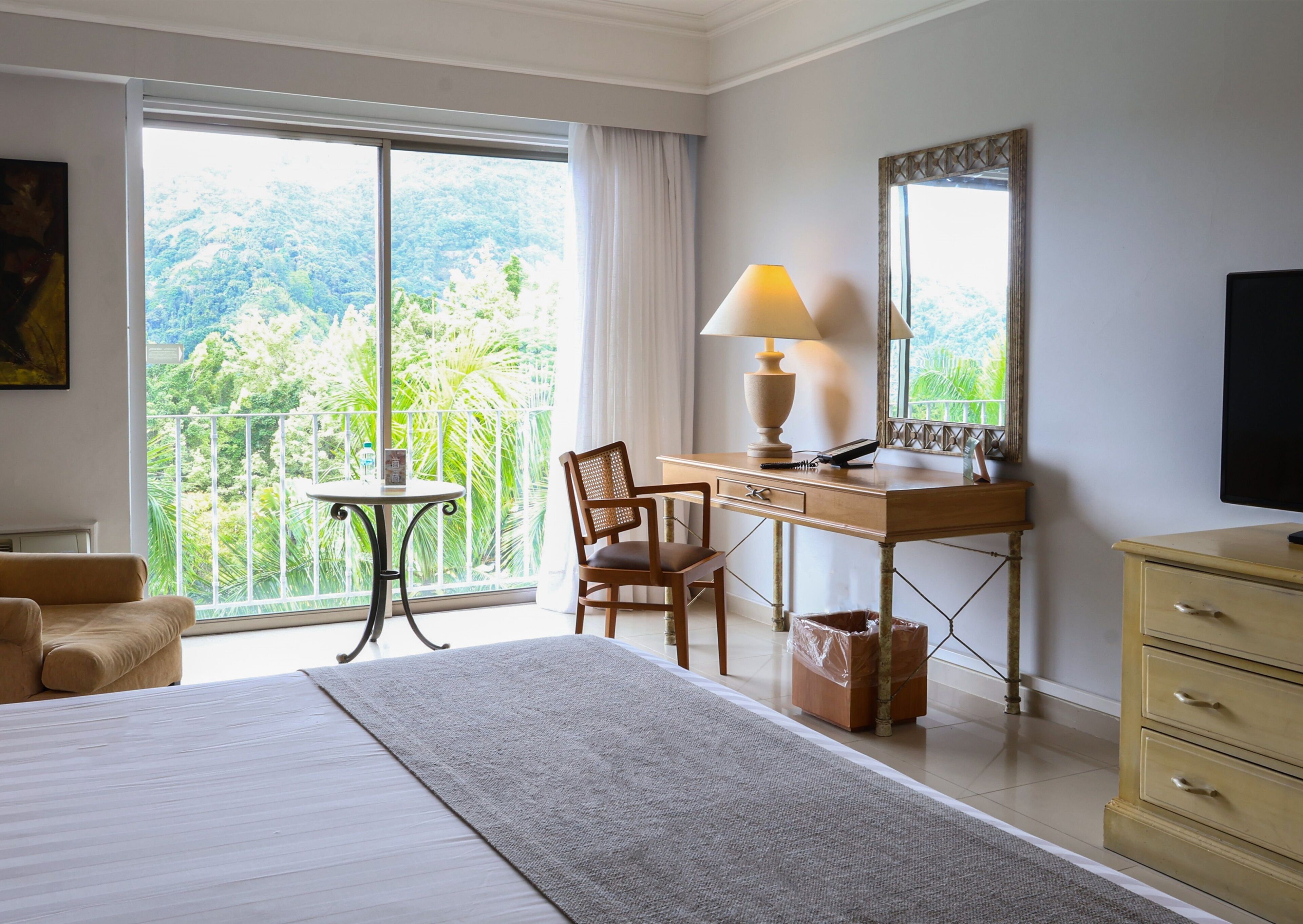 A hotel room with a bed, a desk, a chair, a television, and a balcony with a view of the forest.