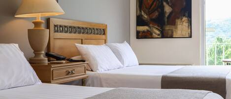 Habitación de hotel con dos camas, una mesita de noche con lámpara, un cuadro en la pared y una ventana con vistas.