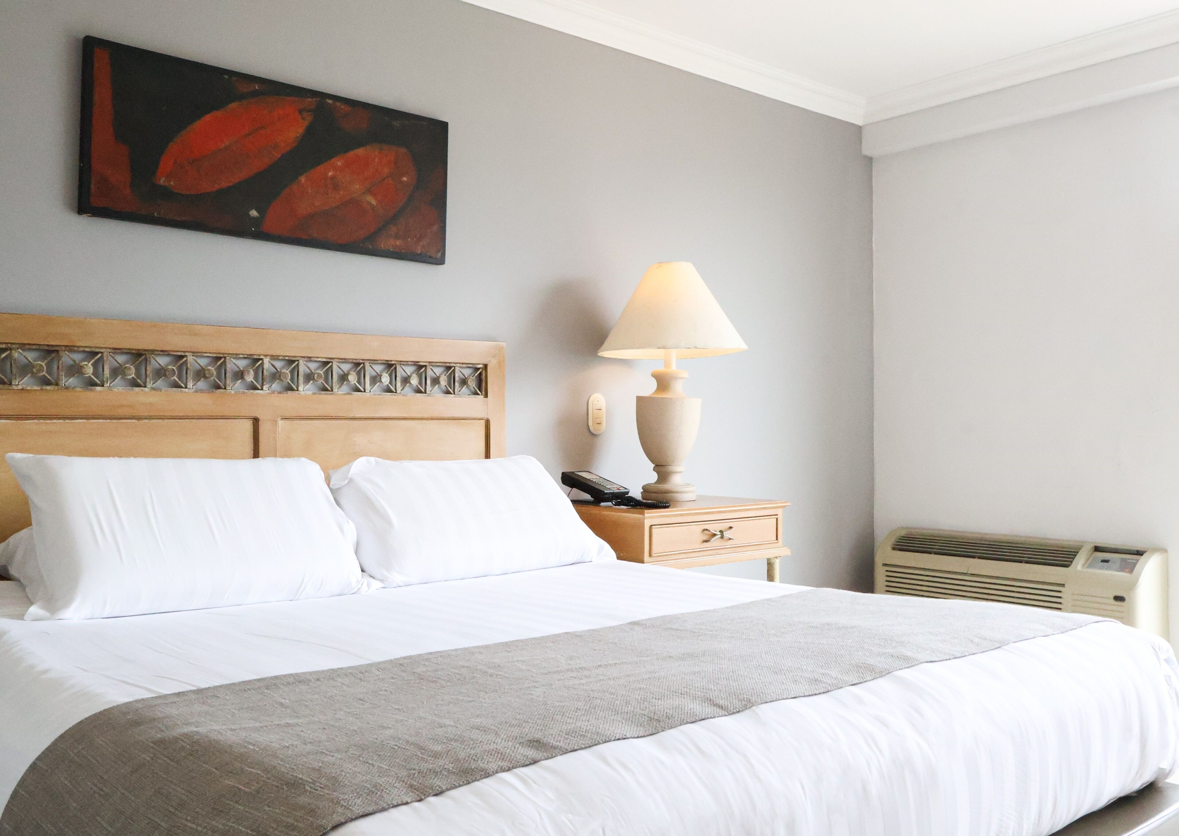 A neatly made bed with white linens and a grey throw blanket, a wooden headboard with a decorative pattern, a bedside lamp, and a wall-mounted painting.