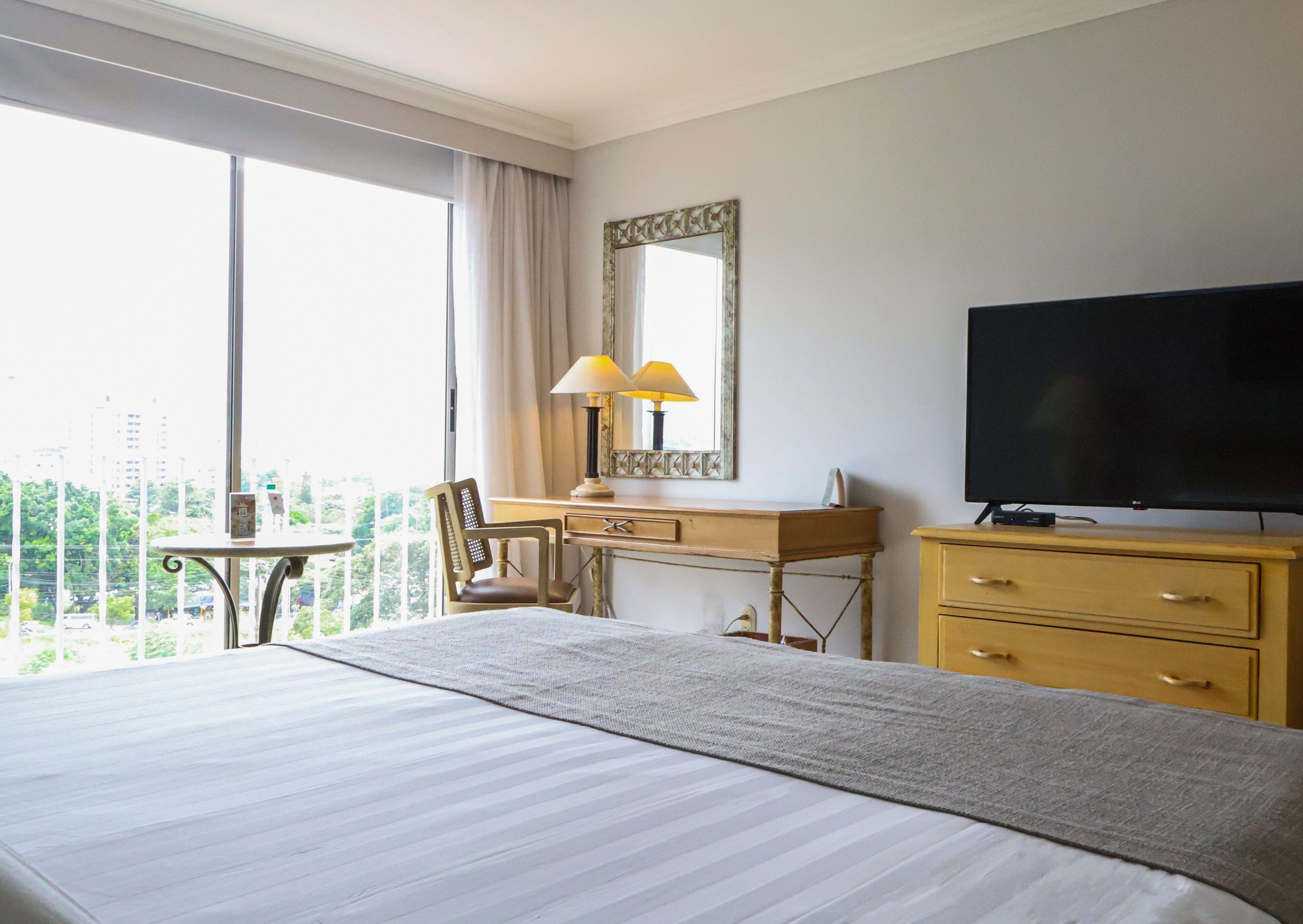 Ein Hotelzimmer mit Bett, Schreibtisch, Stuhl, Fernseher und einem Fenster mit Blick auf Gebäude und Bäume.