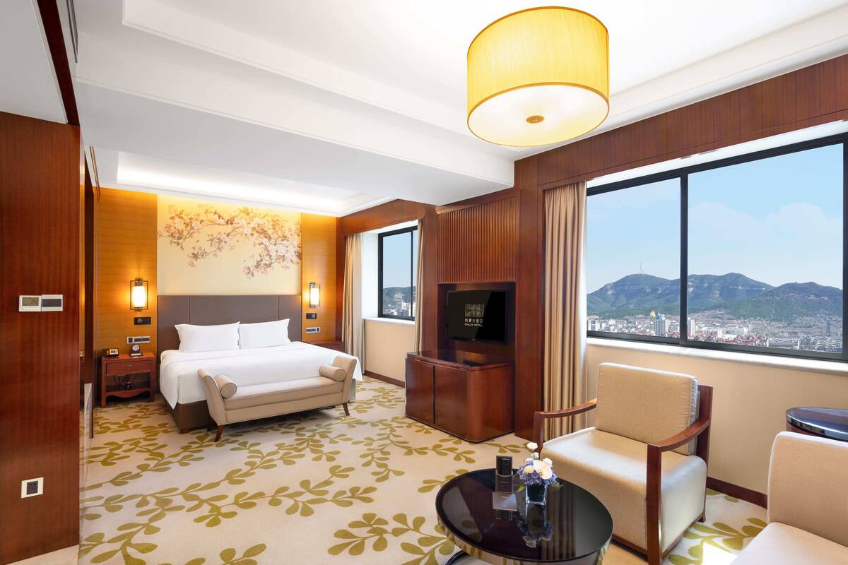 Habitación de hotel con cama, televisor, zona de estar y ventana grande con vista a la ciudad y a las montañas.
