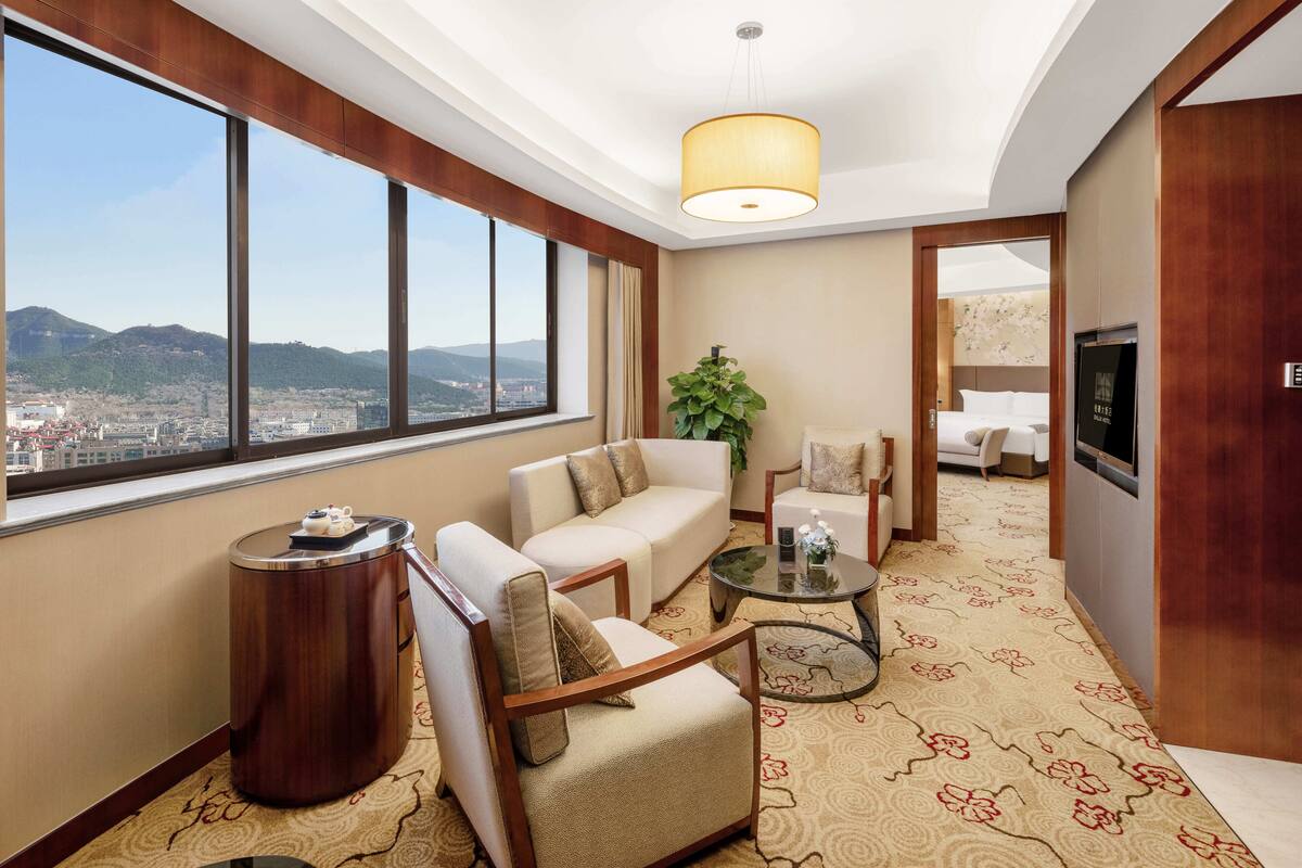 Habitación de hotel moderna con un ventanal grande que ofrece vistas a la ciudad, un sofá, una silla, una mesita y una planta.