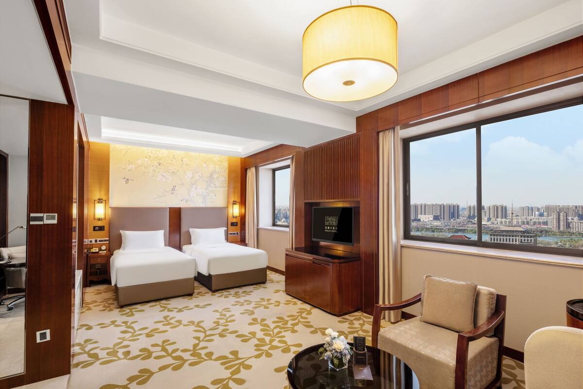 Habitación de hotel con dos camas, televisión, escritorio y una ventana grande con vista a la ciudad.