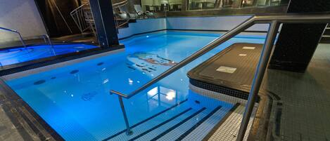 En indendørs swimmingpool med krystalklart vand, flisegulv og gelænder i metal.