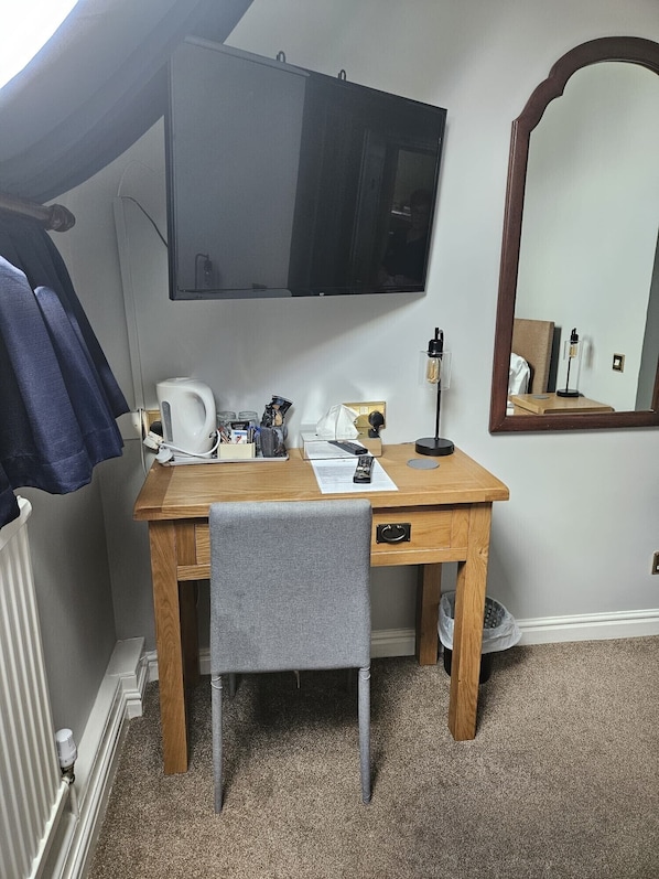 Una habitación pequeña y ordenada con un escritorio de madera, una silla, un televisor de pantalla plana montado en la pared, un espejo y un radiador.
