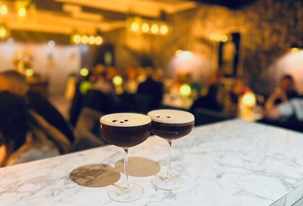 Two glasses of dark liquid on a marble surface with a blurred background of people and lights. - The Oaklands Hotel (Norwich)