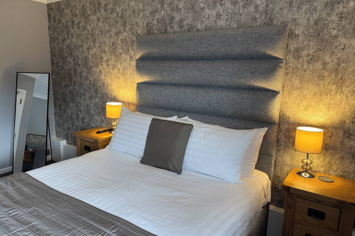 A neatly made bed with white linens and a grey headboard, flanked by two bedside tables with lamps, in a room with textured wallpaper.
