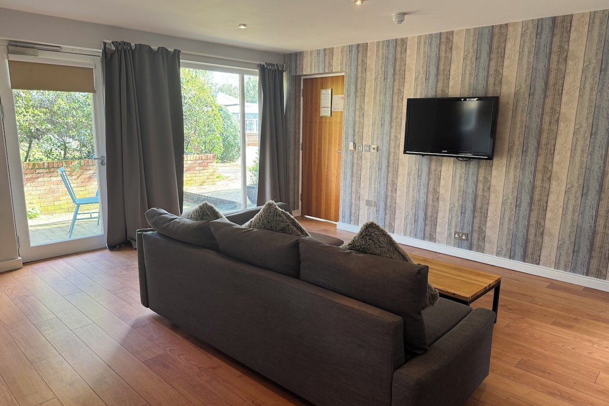 A modern living room with a large sofa, a flat-screen TV mounted on the wall, and a sliding glass door leading to an outdoor area.