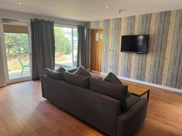 A modern living room with a large sofa, a flat-screen TV mounted on the wall, and a sliding glass door leading to an outdoor area.