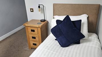 A neatly made bed with a striped bedspread and two pillows, a wooden nightstand with a lamp, and a plain wall.