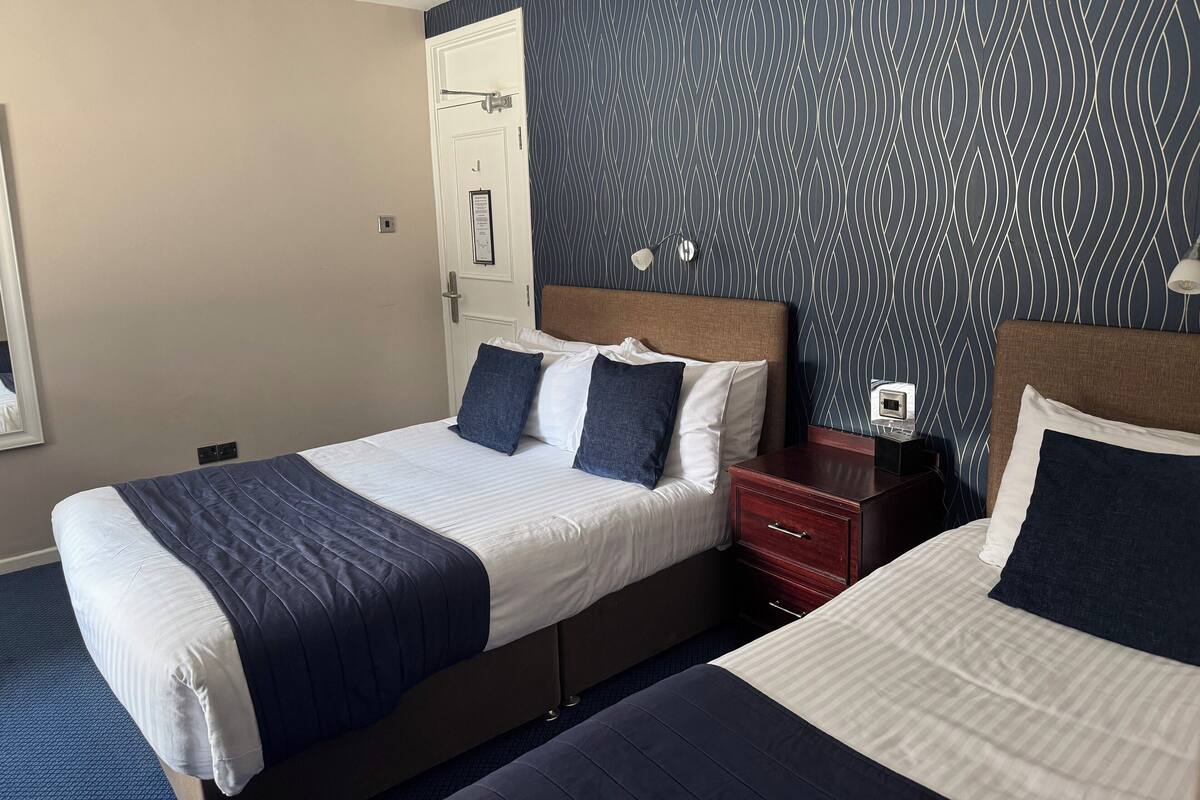 A hotel room with two beds, a patterned wall, a door, and a bedside table with a lamp.