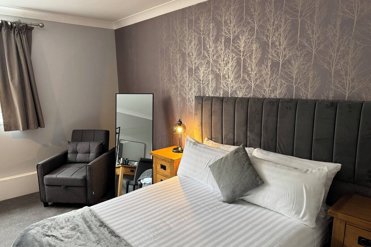 A hotel room with a bed, a chair, a nightstand, a lamp, and a mirror.
