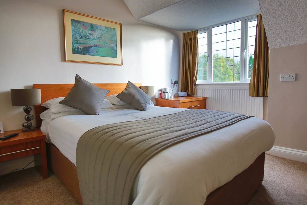 A neatly made bed with a headboard, a bedside table with a lamp, a nightstand, a window with curtains, and a painting on the wall.