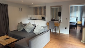A modern living room with a grey sofa, wooden coffee table, and a small desk with a chair. A kitchen area is visible in the background with a wooden cabinet and a mirror. - The Oaklands Hotel (Norwich)