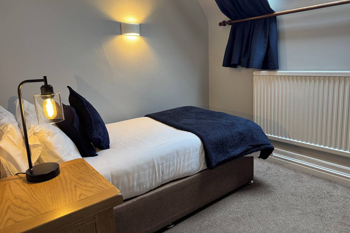 A bedroom with a bed, a bedside table with a lamp, a wall-mounted light, and a window with curtains.