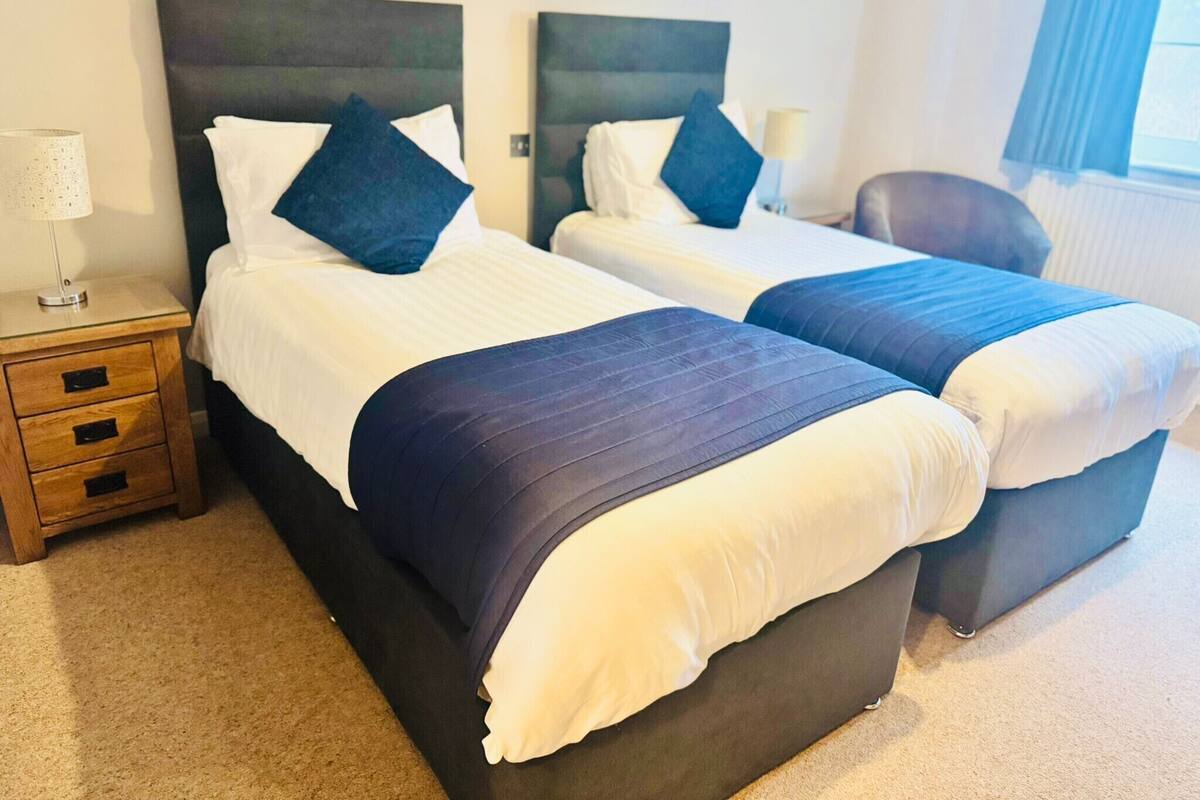 Two single beds with dark headboards and white linens, a wooden nightstand with drawers, a lamp, and a chair in a hotel room.