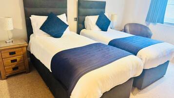 Two single beds with dark headboards and white linens, a wooden nightstand with drawers, a lamp, and a chair in a hotel room.