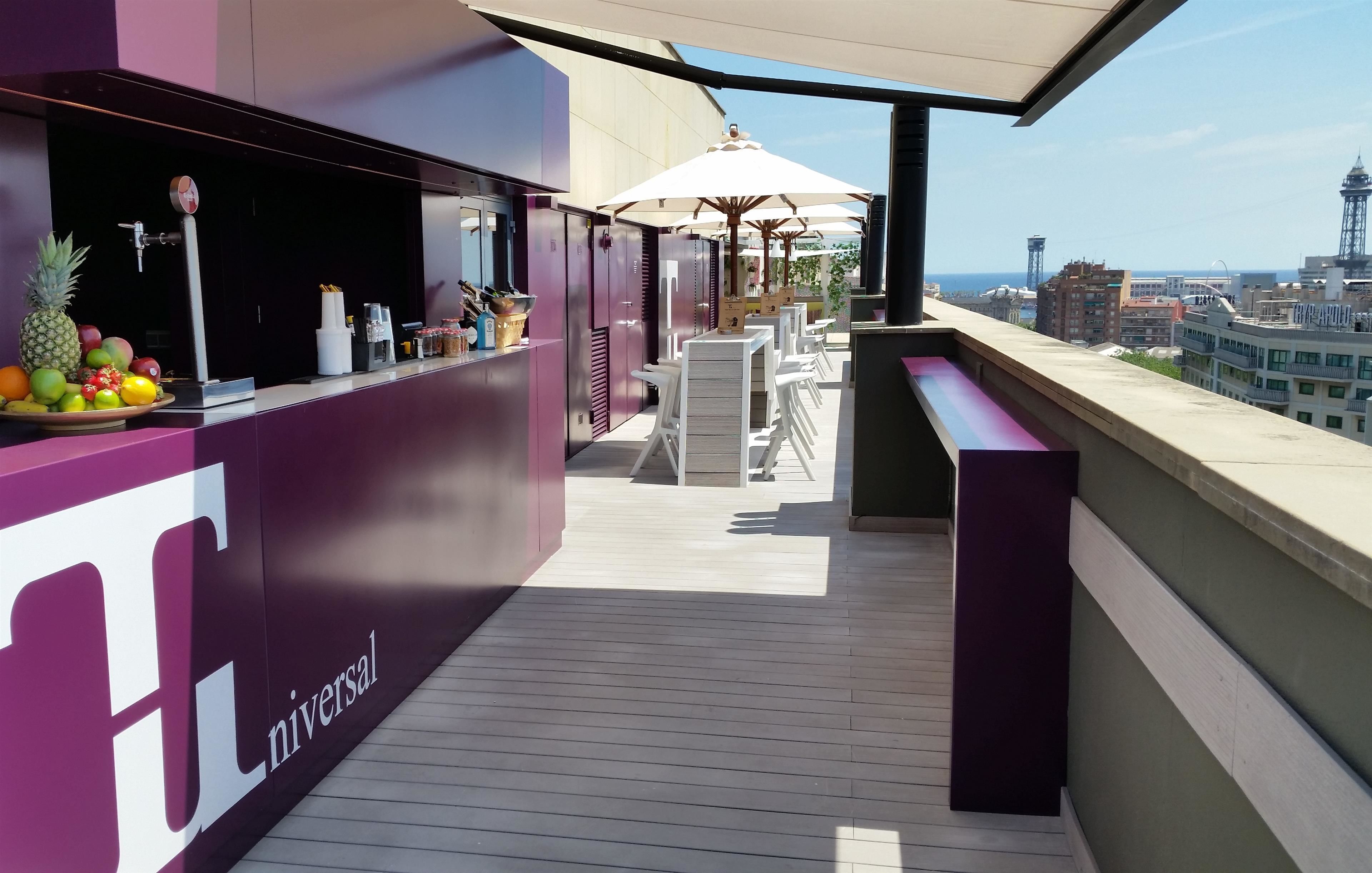 A rooftop bar with a view of the city skyline, featuring a bar counter with a fruit display and outdoor seating.
