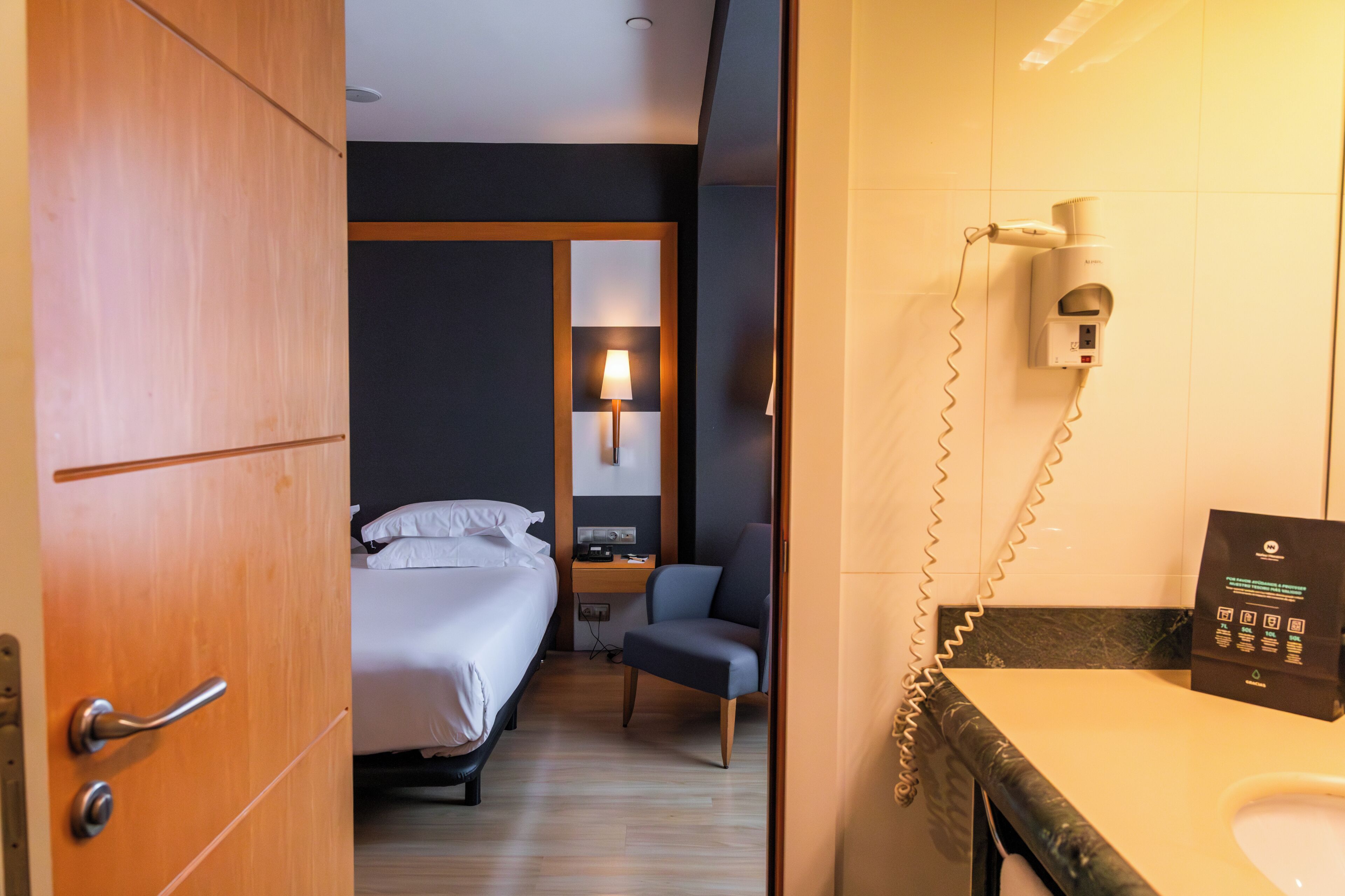 A hotel room with a bed, a desk with a lamp, a chair, a hairdryer, and a bathroom with a sink and mirror.